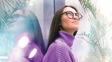 A woman in glasses and a purple jumper stands with her back to a reflective surface 