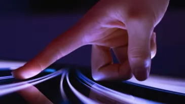 An extended finger points downward to a shiny digital surface, where multiple contours of light display.